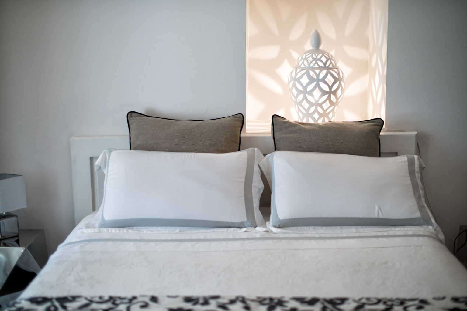 Cozy bedroom setup featuring a neatly made bed with white pillows and decorative beige cushions, complemented by an elegant lamp casting soft light through a patterned window. | Only on SopranoVillas