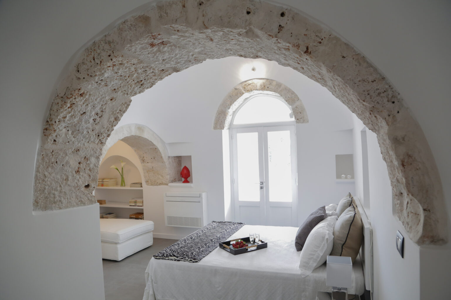 Cozy bedroom with white walls and traditional stone archway, featuring a comfortable bed, modern decor, and natural light streaming through large windows. | Only on SopranoVillas