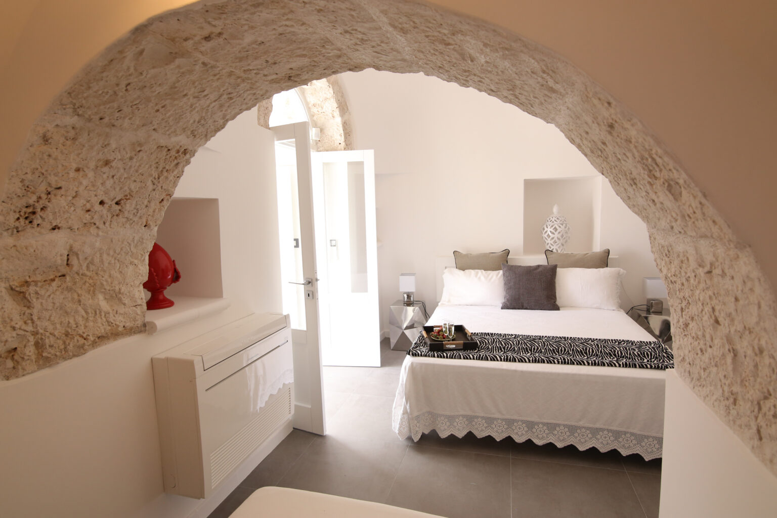 Spacious bedroom featuring a comfortable bed with decorative pillows, a minimalist dresser, and an elegant archway leading to a bright entrance. The room is designed with neutral tones and modern decor, creating a serene atmosphere. | Only on SopranoVillas
