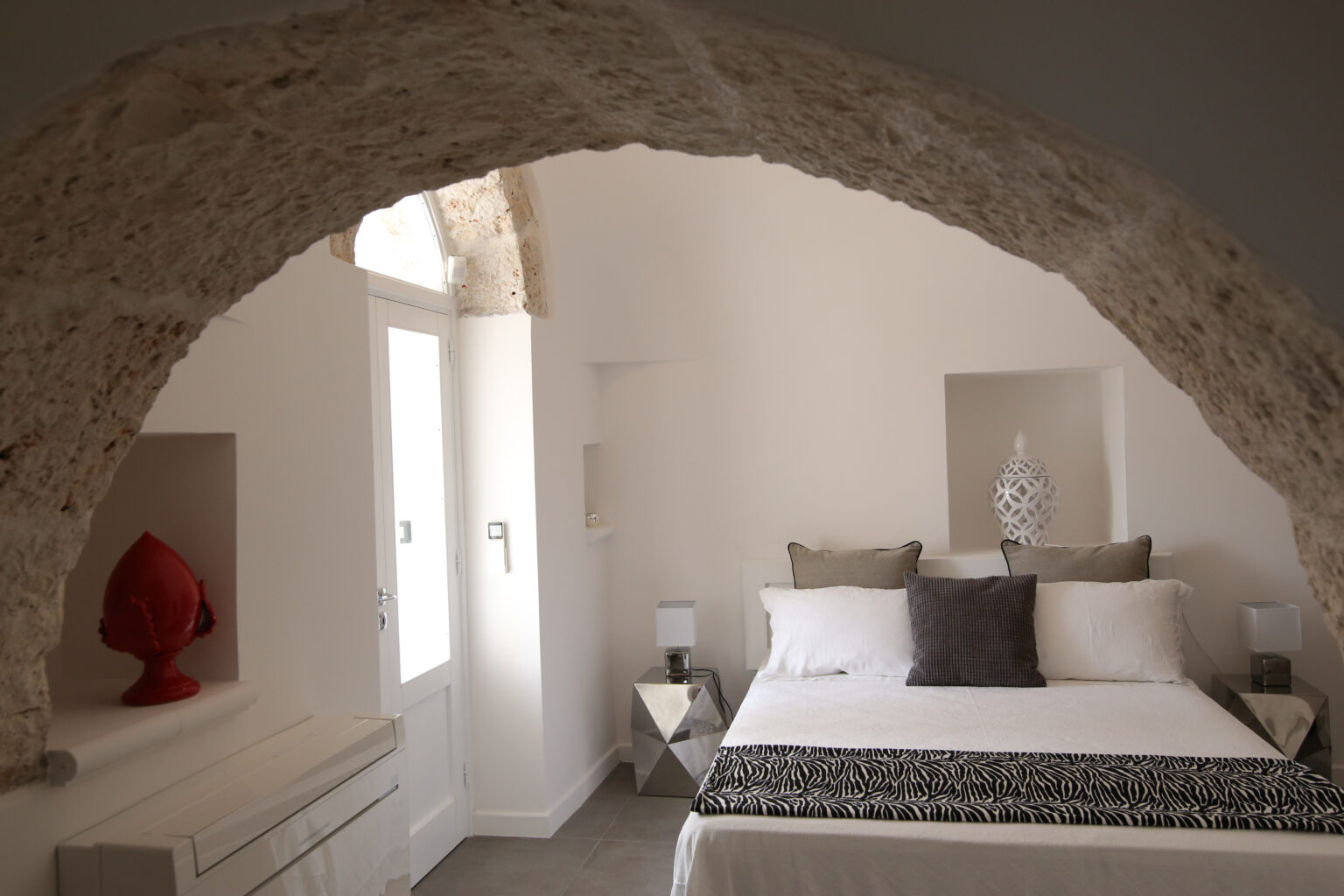 Cozy bedroom featuring a modern design with a large bed, decorative pillows, and natural light streaming through arched windows. The room has a stylish, minimalist aesthetic with stone walls and contemporary furnishings. | Only on SopranoVillas