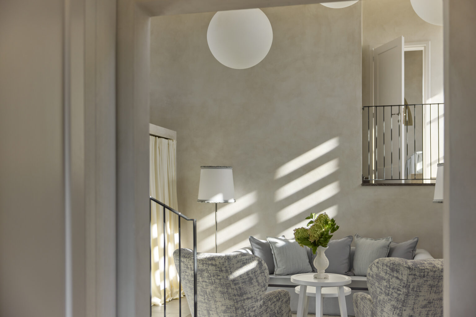 Cozy modern living room with soft gray furnishings, a round table, and a vase of green flowers, illuminated by natural light through large windows and a pendant light fixture. | Only on SopranoVillas