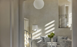 Cozy modern living room with soft gray furnishings, a round table, and a vase of green flowers, illuminated by natural light through large windows and a pendant light fixture.