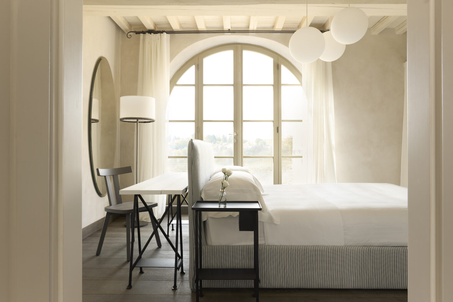 Modern bedroom interior featuring a large arched window, minimalist furniture including a bed, desk, and chair, soft natural light, and decorative pendant lamps for a cozy ambiance. | Only on SopranoVillas
