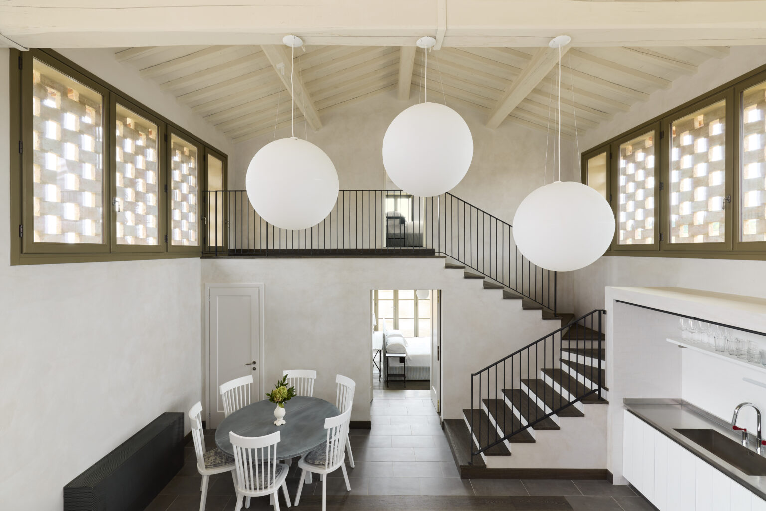 Modern interior design featuring a spacious living area with a round dining table, white chairs, and large pendant lights. The room includes a staircase with a sleek railing and decorative windows that allow natural light to illuminate the space. | Only on SopranoVillas
