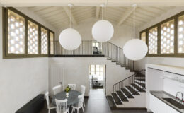 Modern interior design featuring a spacious living area with a round dining table, white chairs, and large pendant lights. The room includes a staircase with a sleek railing and decorative windows that allow natural light to illuminate the space.