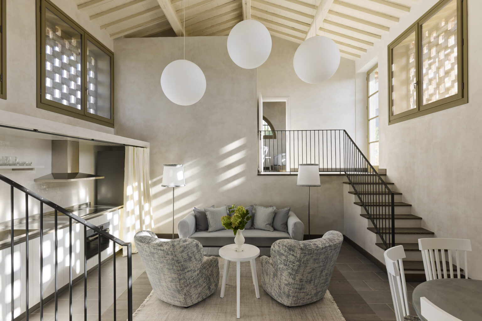 Modern living room with a cozy seating area featuring plush gray chairs and a white coffee table, illuminated by three spherical pendant lights. The open layout includes a staircase leading to a second floor, with large windows allowing natural light to fill the space. The neutral color palette creates a serene atmosphere, ideal for contemporary home design. | Only on SopranoVillas