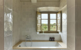 Modern bathroom featuring a bathtub with a natural stone countertop, open wooden windows providing a scenic view, and neatly arranged toiletries on the ledge.