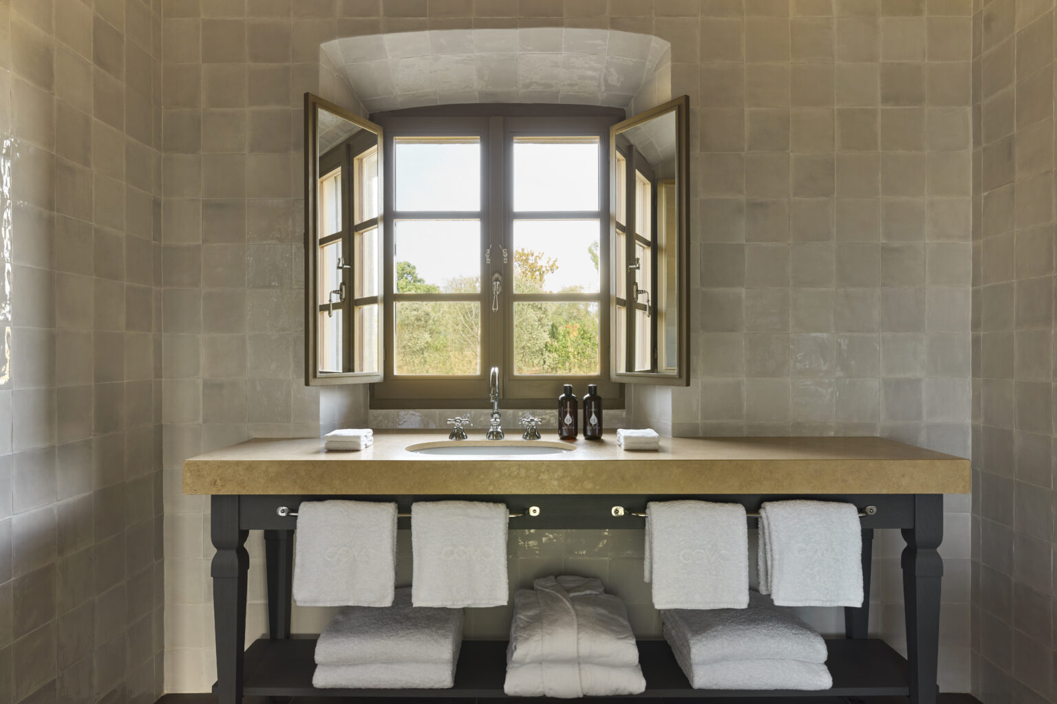 Elegant bathroom featuring a wooden vanity with a double sink, neatly arranged towels, and large windows allowing natural light to illuminate the stone walls and serene outdoor view. | Only on SopranoVillas