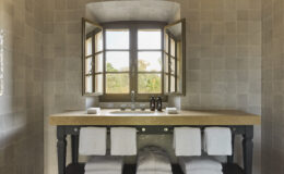 Elegant bathroom featuring a wooden vanity with a double sink, neatly arranged towels, and large windows allowing natural light to illuminate the stone walls and serene outdoor view.