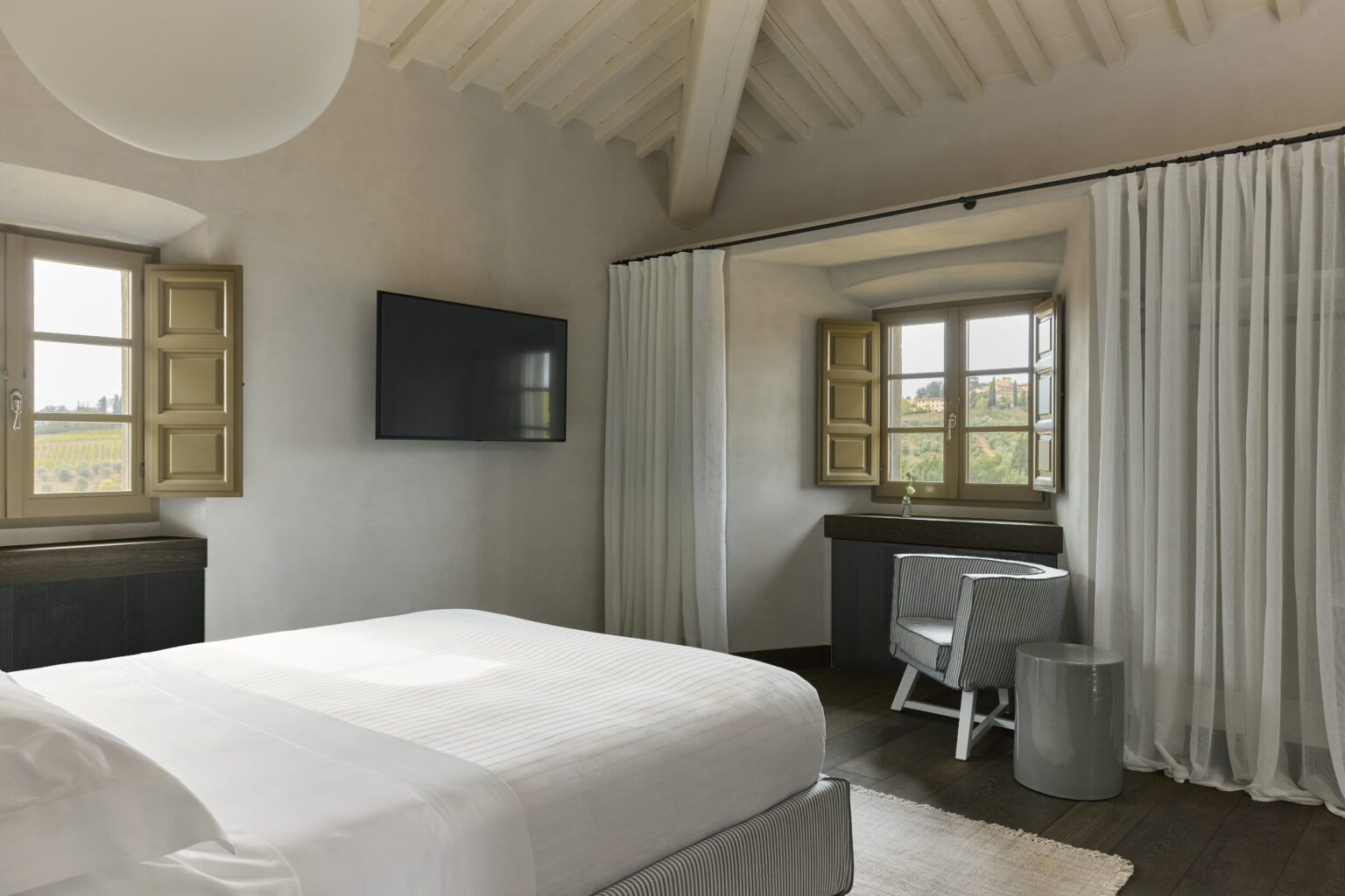 Modern bedroom interior featuring a spacious white bed, a wall-mounted television, and large windows with wooden shutters. The room is accented with soft curtains and minimalist furniture, creating a bright and airy atmosphere. | Only on SopranoVillas
