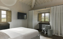 Modern bedroom interior featuring a spacious white bed, a wall-mounted television, and large windows with wooden shutters. The room is accented with soft curtains and minimalist furniture, creating a bright and airy atmosphere.