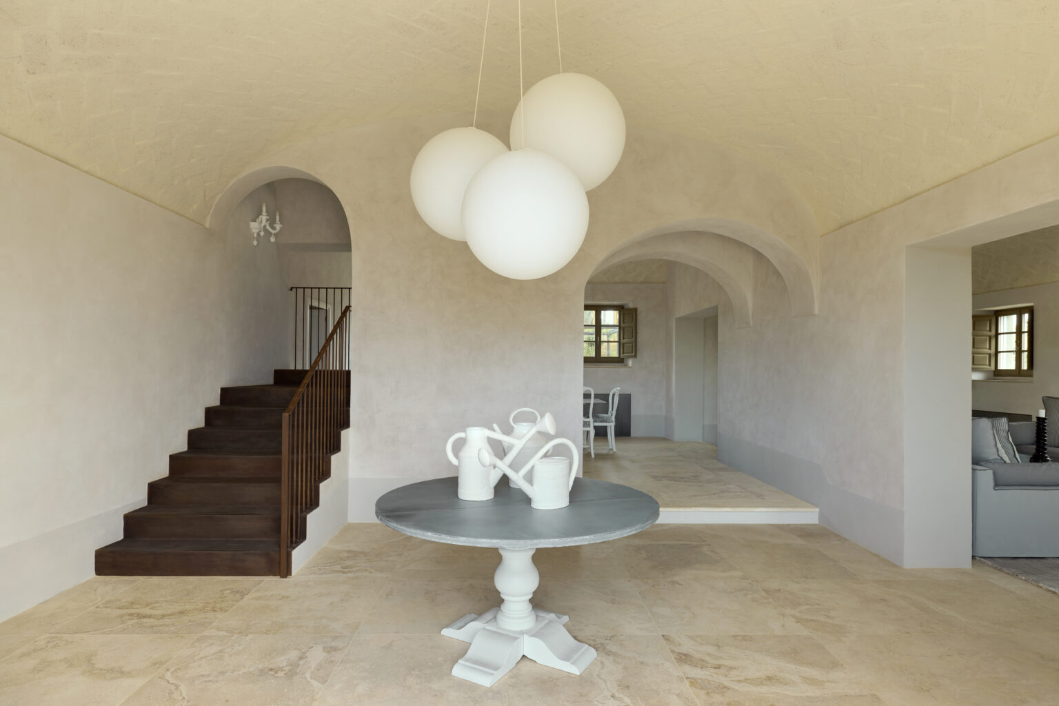 Modern interior design featuring a spacious living area with a round table and decorative vases, complemented by a minimalist staircase and stylish pendant lighting. Natural light enhances the warm tones of the walls and flooring, creating an inviting atmosphere. | Only on SopranoVillas