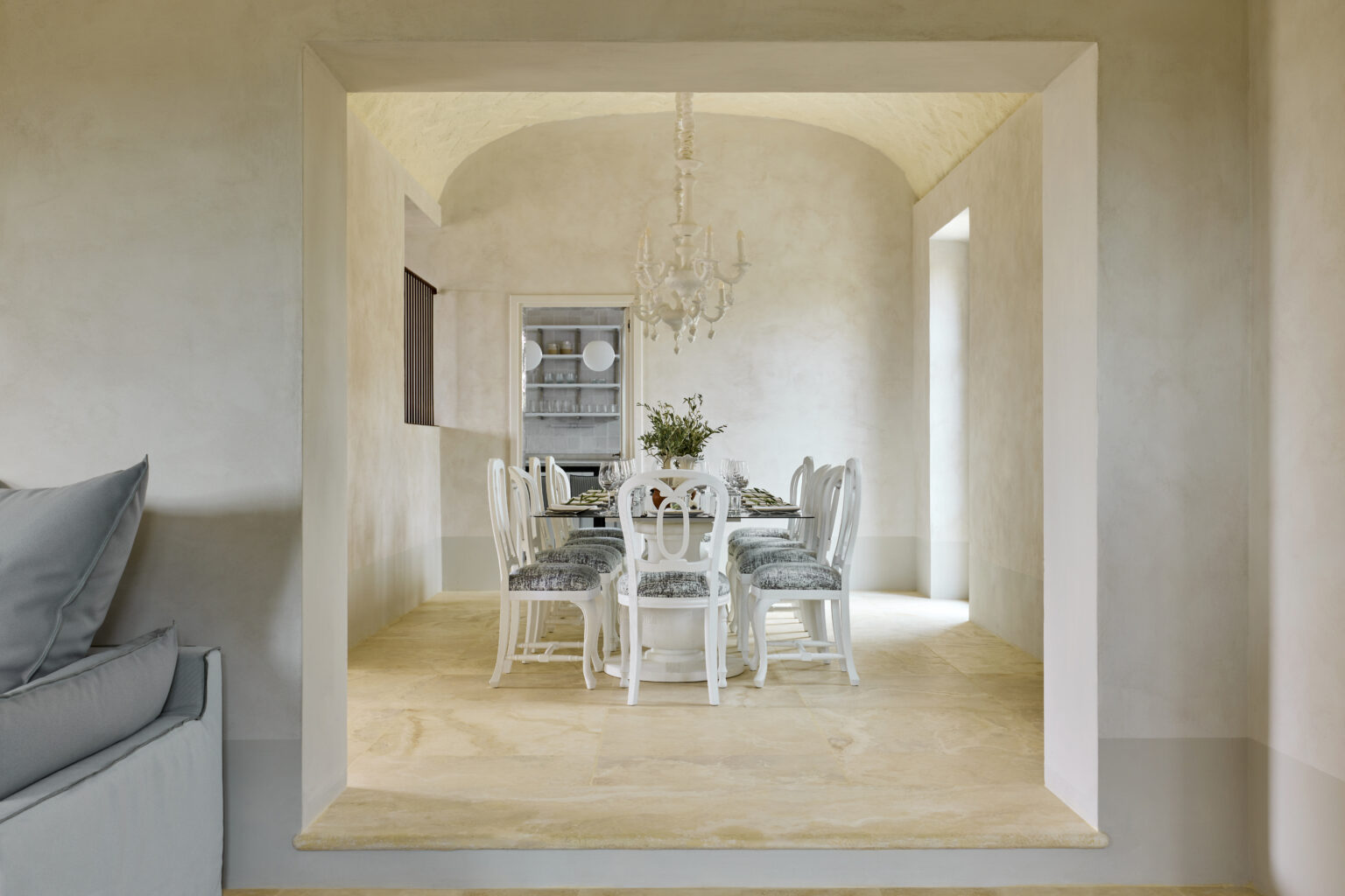 Elegant dining room featuring a glass table surrounded by white chairs with gray upholstery, illuminated by a stylish chandelier. The room showcases a soft color palette with textured walls and a warm stone floor, creating a sophisticated atmosphere. | Only on SopranoVillas