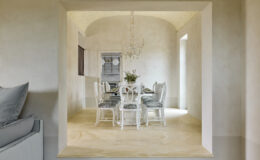 Elegant dining room featuring a glass table surrounded by white chairs with gray upholstery, illuminated by a stylish chandelier. The room showcases a soft color palette with textured walls and a warm stone floor, creating a sophisticated atmosphere.