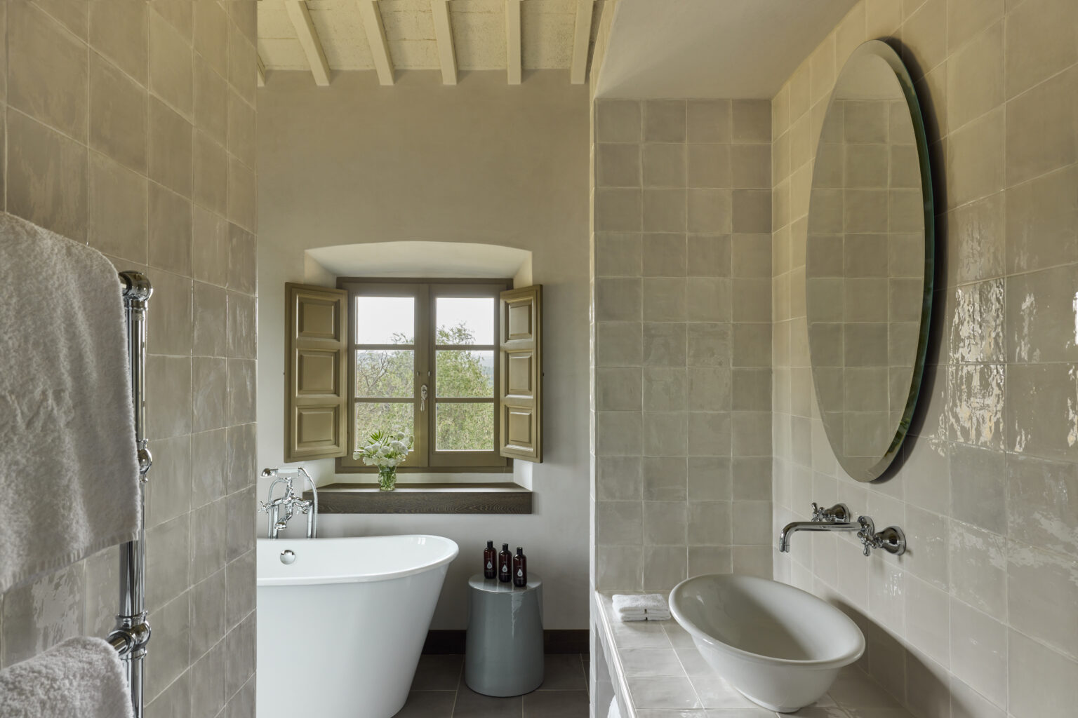 Modern bathroom design featuring a freestanding bathtub, a sleek sink, and large windows with wooden shutters, enhancing natural light and providing a serene view. The neutral tile and decor create a calming atmosphere. | Only on SopranoVillas