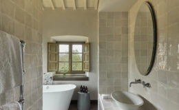 Modern bathroom design featuring a freestanding bathtub, a sleek sink, and large windows with wooden shutters, enhancing natural light and providing a serene view. The neutral tile and decor create a calming atmosphere.