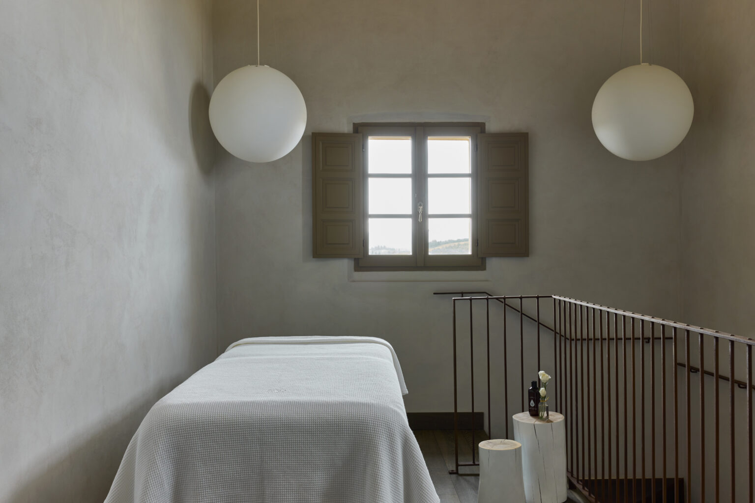 Minimalist spa room featuring a massage table, soft lighting from spherical pendant lamps, and a window with wooden shutters, creating a serene atmosphere for relaxation and wellness. | Only on SopranoVillas