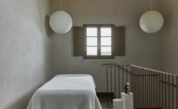 Minimalist spa room featuring a massage table, soft lighting from spherical pendant lamps, and a window with wooden shutters, creating a serene atmosphere for relaxation and wellness.