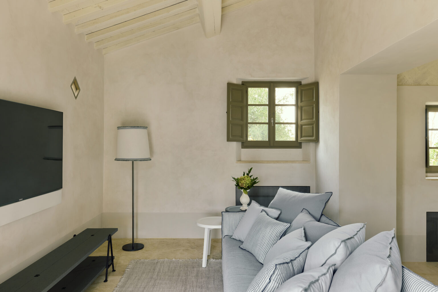 Modern living room featuring a light color palette, a comfortable gray sofa adorned with decorative pillows, a sleek black TV mounted on the wall, and a stylish floor lamp. Natural light filters through green-shuttered windows, enhancing the inviting atmosphere. | Only on SopranoVillas
