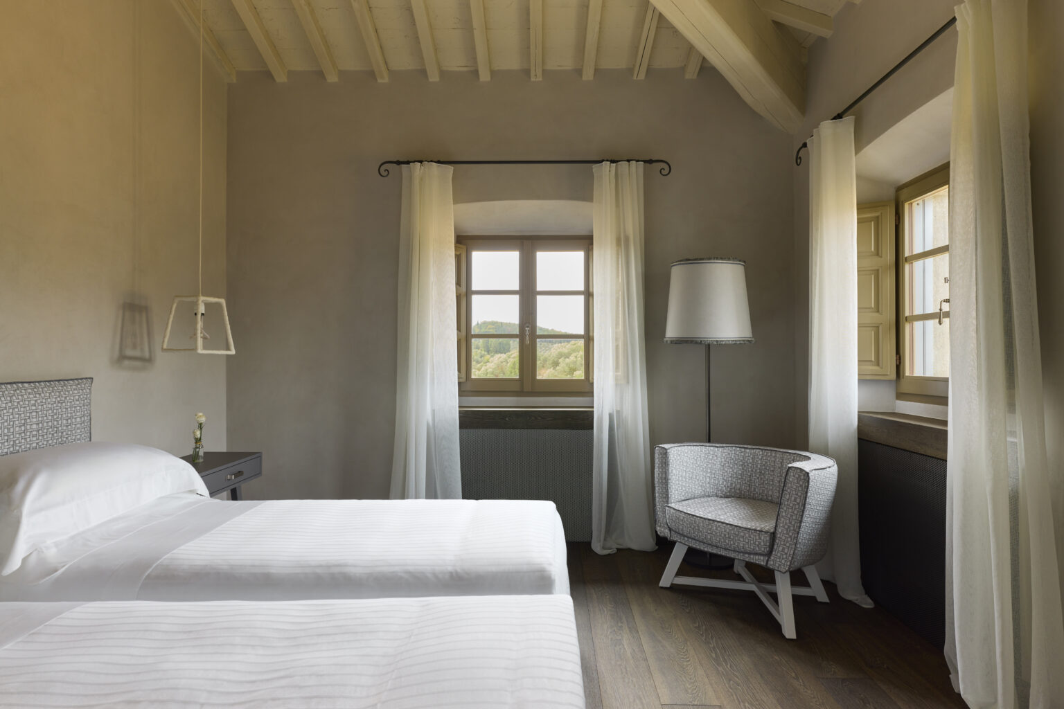 Cozy bedroom featuring two twin beds, a modern armchair, and a large window with natural light, complemented by soft curtains and rustic wooden flooring. | Only on SopranoVillas