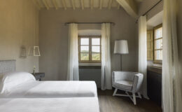 Cozy bedroom featuring two twin beds, a modern armchair, and a large window with natural light, complemented by soft curtains and rustic wooden flooring.