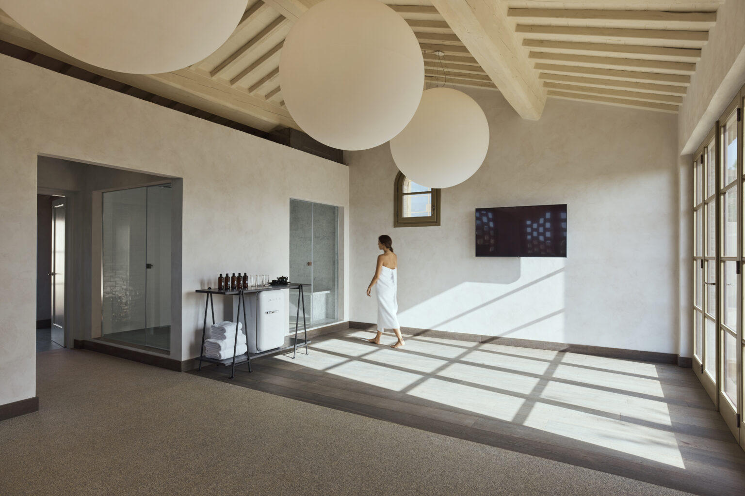 Modern spa interior with natural light, featuring a woman in a white robe walking on a polished floor, large round pendant lights, and glass doors. Ideal for wellness and relaxation themes. | Only on SopranoVillas