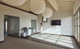 Modern spa interior with natural light, featuring a woman in a white robe walking on a polished floor, large round pendant lights, and glass doors. Ideal for wellness and relaxation themes.