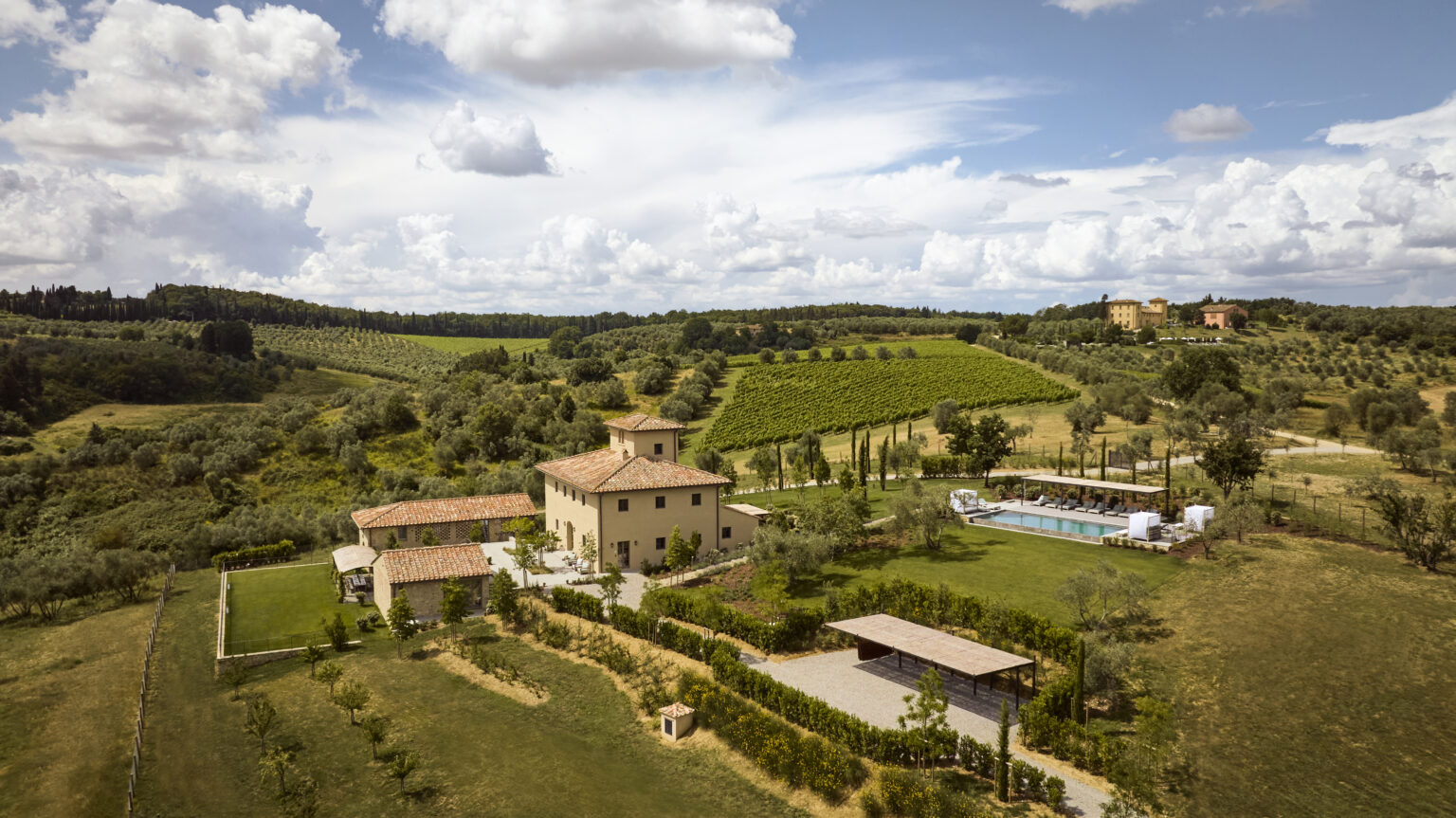 Aerial view of a picturesque Italian villa surrounded by lush green vineyards and rolling hills, featuring a swimming pool and landscaped gardens under a partly cloudy sky. | Only on SopranoVillas