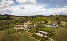Aerial view of a picturesque Italian villa surrounded by lush green vineyards and rolling hills, featuring a swimming pool and landscaped gardens under a partly cloudy sky.