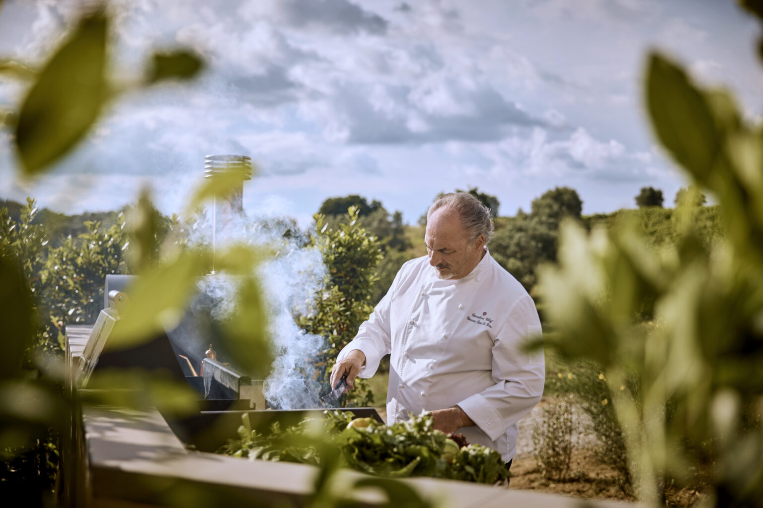 Chef grilling outdoors with smoke rising, surrounded by greenery and a scenic landscape. The chef wears a white culinary jacket and focuses on preparing fresh ingredients on a barbecue grill. Ideal for content related to outdoor cooking, culinary arts, and gourmet recipes. | Only on SopranoVillas