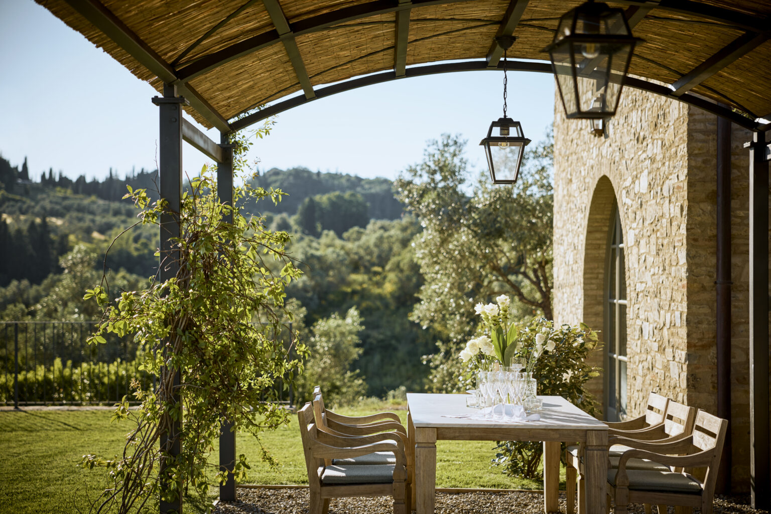 Outdoor dining area with a wooden table and chairs, surrounded by lush greenery and a scenic view of rolling hills, featuring a stone building and hanging lanterns under a thatched roof. Perfect for enjoying meals in a tranquil setting. | Only on SopranoVillas