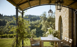 Outdoor dining area with a wooden table and chairs, surrounded by lush greenery and a scenic view of rolling hills, featuring a stone building and hanging lanterns under a thatched roof. Perfect for enjoying meals in a tranquil setting.