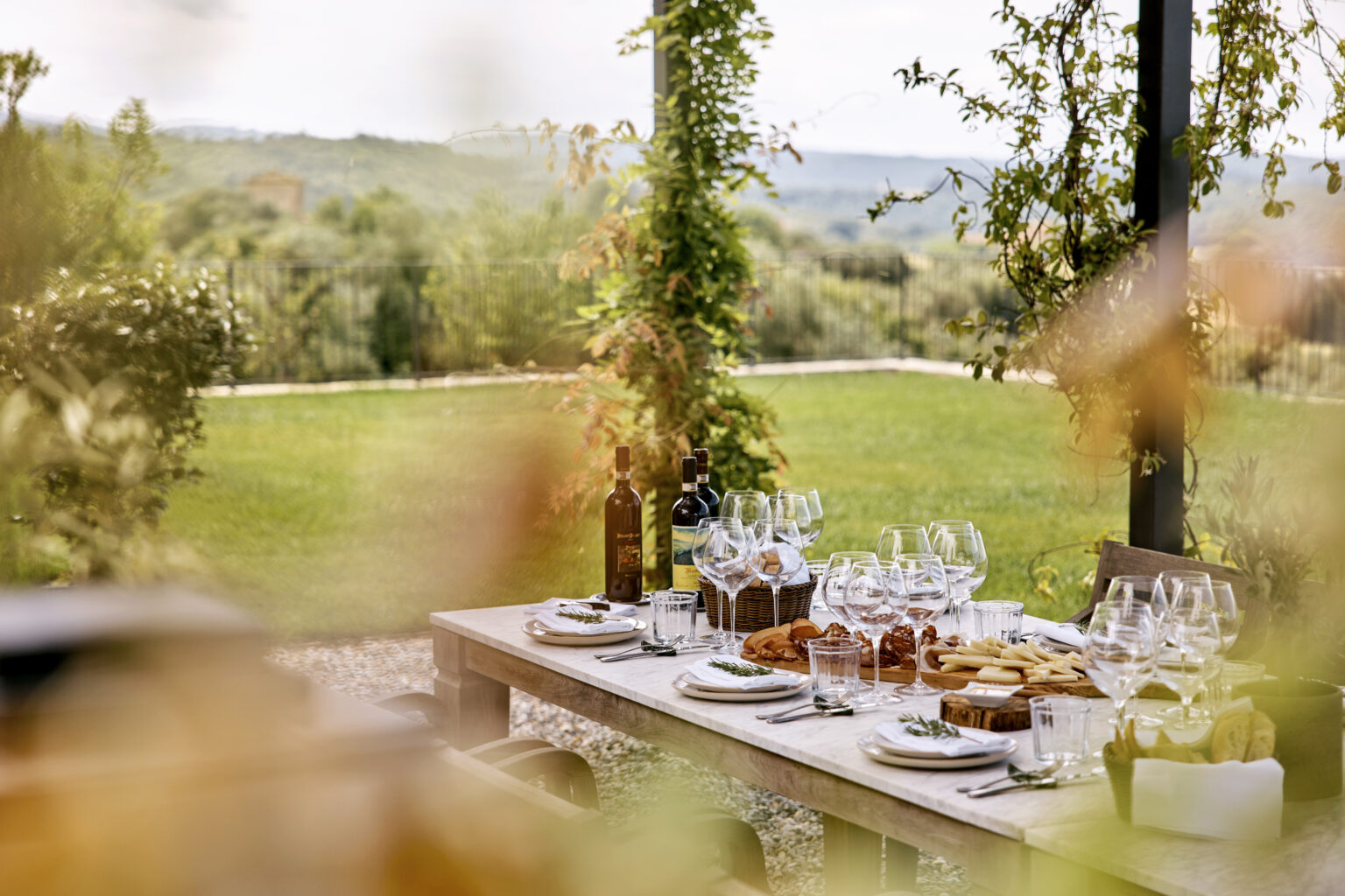 Outdoor dining setup featuring a wooden table adorned with elegant glassware, plates of food, and bottles of wine, set against a lush green landscape and distant hills. Perfect for a gourmet meal or gathering in a scenic setting. | Only on SopranoVillas