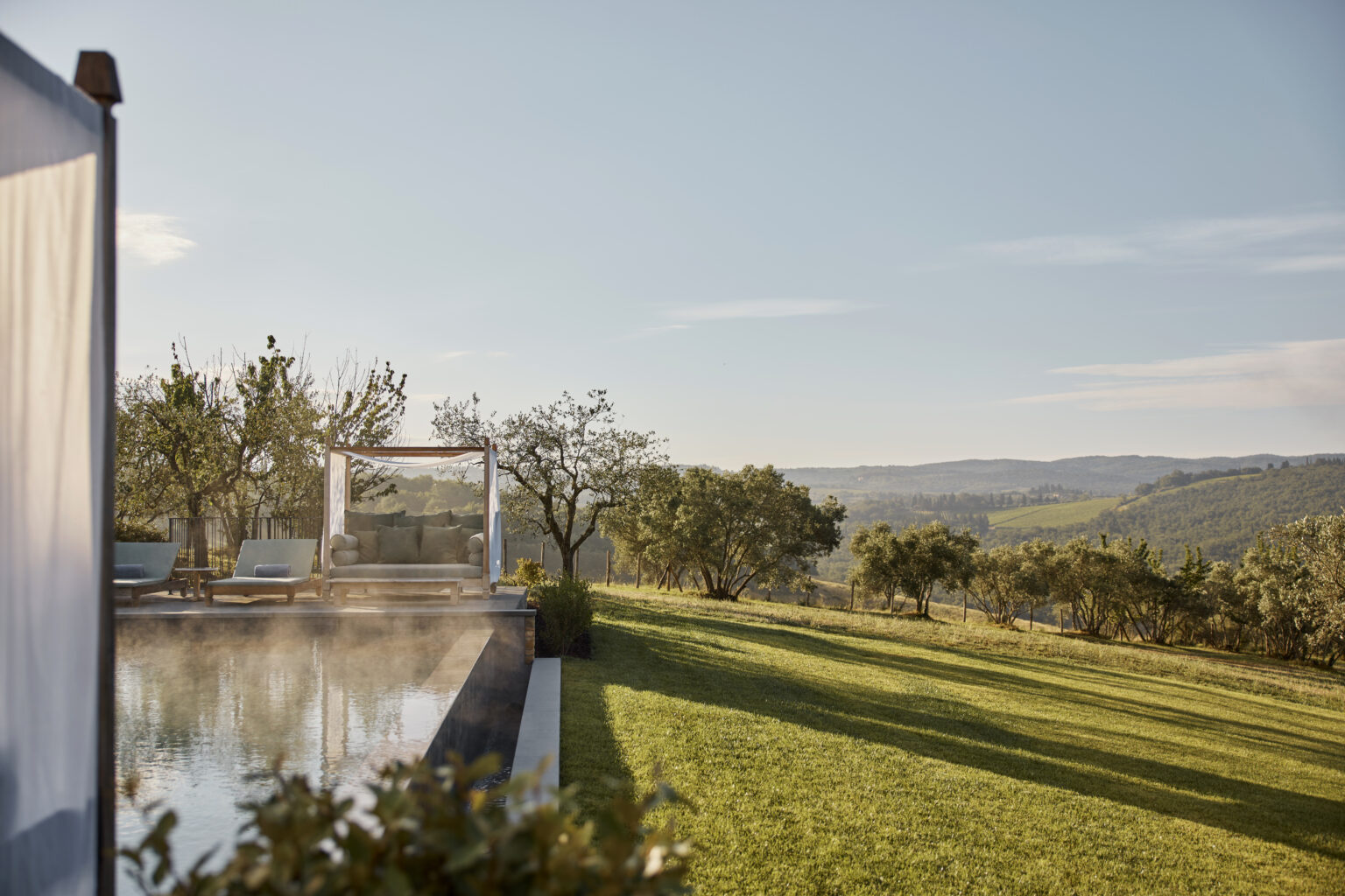 Scenic view of a tranquil outdoor space featuring a swimming pool, comfortable lounge area with a shaded canopy, and lush green lawn, set against rolling hills and clear blue skies. Ideal for relaxation and enjoying nature. | Only on SopranoVillas