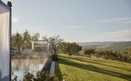 Scenic view of a tranquil outdoor space featuring a swimming pool, comfortable lounge area with a shaded canopy, and lush green lawn, set against rolling hills and clear blue skies. Ideal for relaxation and enjoying nature.