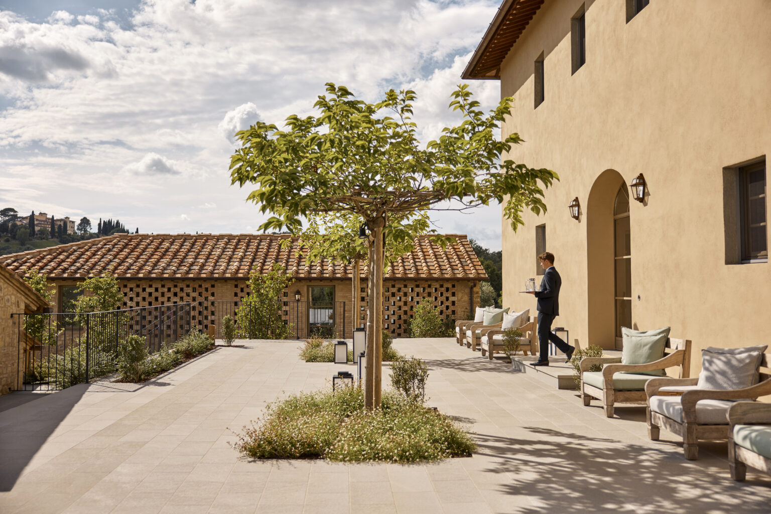 A stylish outdoor terrace at a Tuscan villa, featuring a young man in formal attire carrying a tray, surrounded by greenery, wooden seating, and a rustic tiled roof in the background under a partly cloudy sky. | Only on SopranoVillas