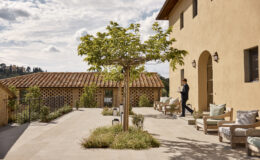 A stylish outdoor terrace at a Tuscan villa, featuring a young man in formal attire carrying a tray, surrounded by greenery, wooden seating, and a rustic tiled roof in the background under a partly cloudy sky.