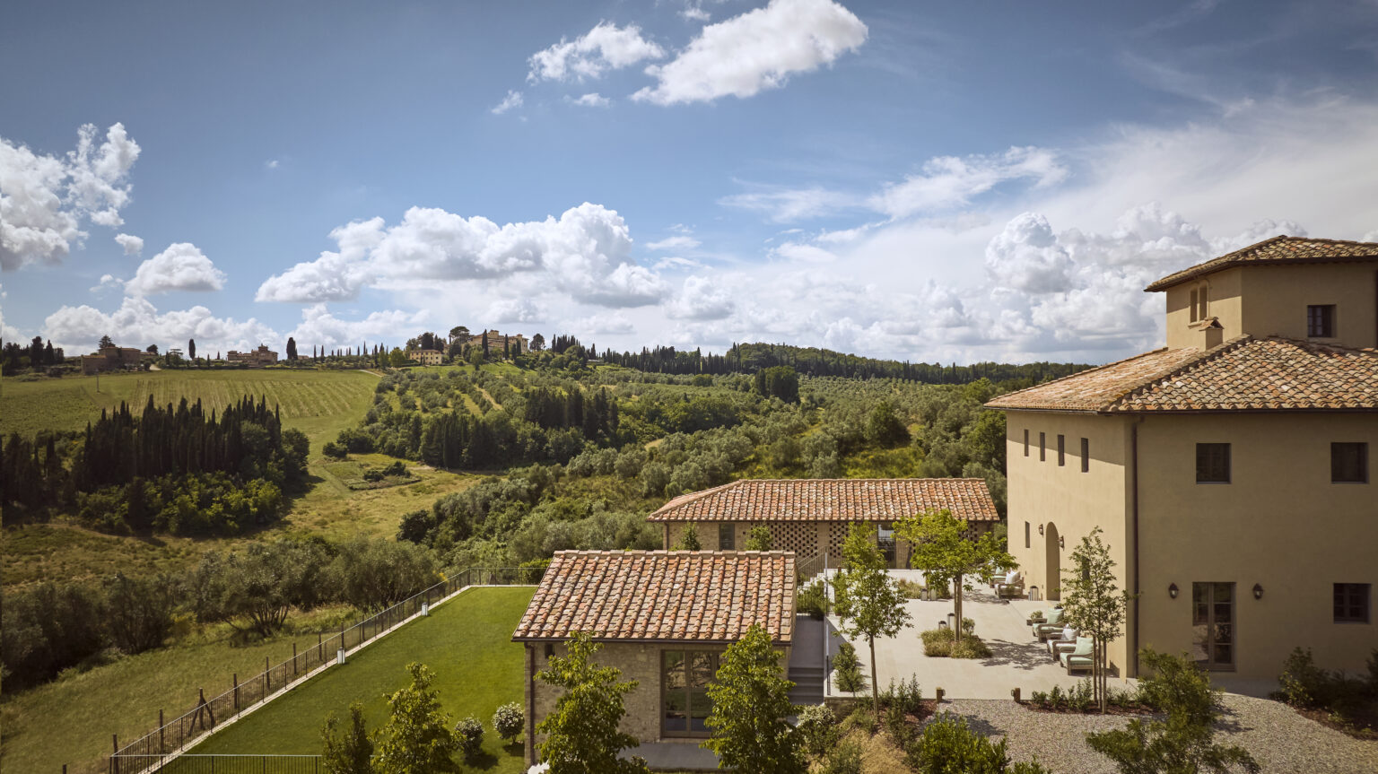 Scenic view of a Tuscan villa surrounded by lush green hills and trees under a partly cloudy sky, showcasing traditional architecture and outdoor spaces. | Only on SopranoVillas