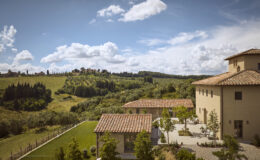 Scenic view of a Tuscan villa surrounded by lush green hills and trees under a partly cloudy sky, showcasing traditional architecture and outdoor spaces.