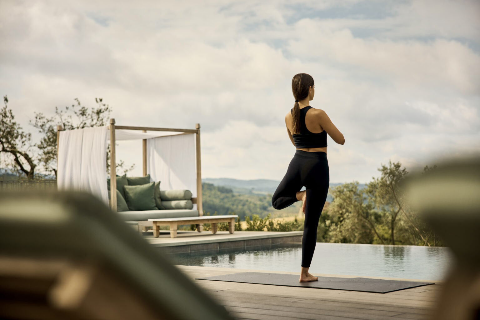 A woman practicing yoga on a mat by an infinity pool, surrounded by scenic hills and a modern outdoor lounge area. The setting promotes relaxation and wellness, ideal for yoga enthusiasts. | Only on SopranoVillas