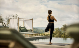 A woman practicing yoga on a mat by an infinity pool, surrounded by scenic hills and a modern outdoor lounge area. The setting promotes relaxation and wellness, ideal for yoga enthusiasts.