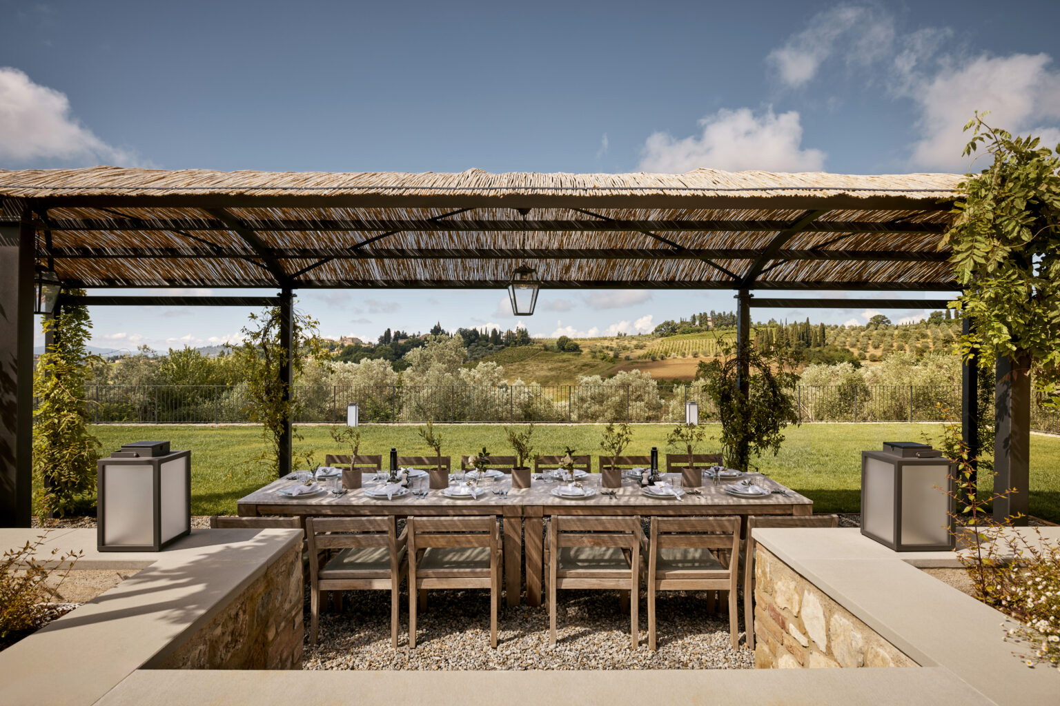 Outdoor dining area under a straw-covered pergola, featuring a long wooden table set for a meal, surrounded by lush greenery and rolling hills in the background. Ideal for alfresco dining experiences in a picturesque landscape. | Only on SopranoVillas