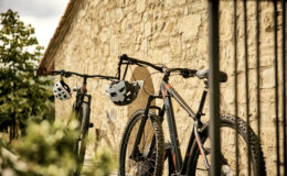 Two mountain bikes with helmets hanging on handlebars, positioned against a rustic stone wall, surrounded by greenery.