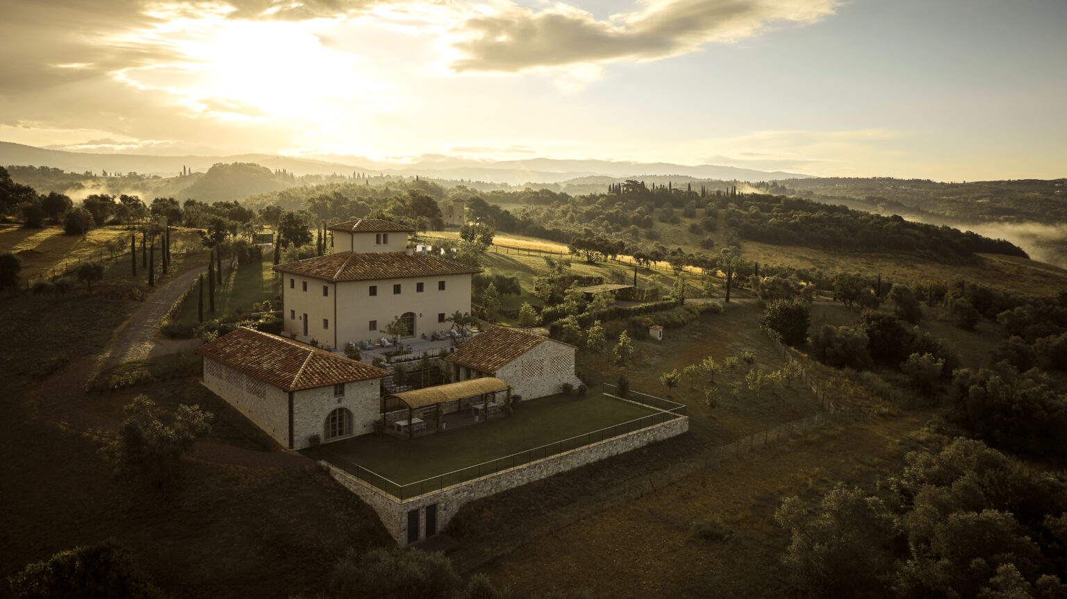 Aerial view of a picturesque Italian villa surrounded by lush green hills and fields at sunset, showcasing the rustic architecture and serene landscape. | Only on SopranoVillas