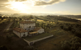 Aerial view of a picturesque Italian villa surrounded by lush green hills and fields at sunset, showcasing the rustic architecture and serene landscape.