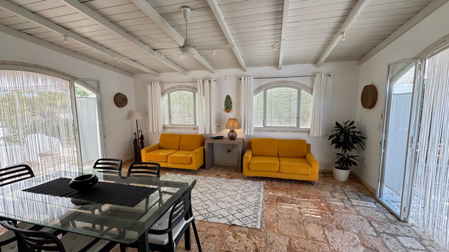 Bright and airy living room featuring two yellow sofas, a glass dining table, and large windows allowing natural light. The space is decorated with indoor plants and has a cozy, inviting atmosphere. | Only on SopranoVillas