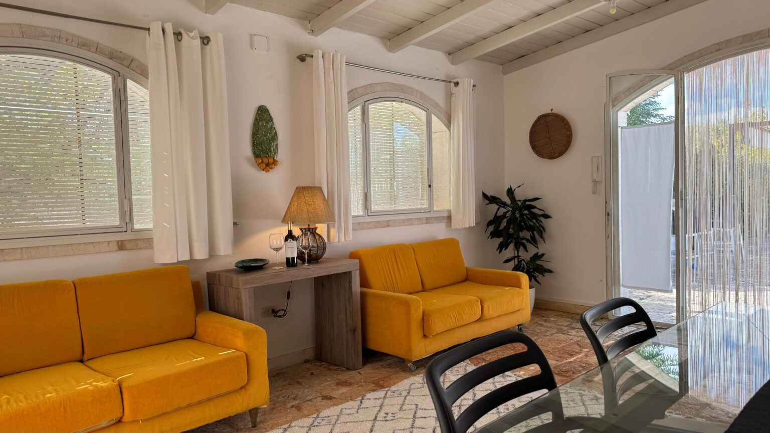 Bright and cozy living room featuring two yellow sofas, a wooden side table with a lamp, a decorative wall piece, and indoor plants, with large windows allowing natural light and a view of an outdoor area. | Only on SopranoVillas