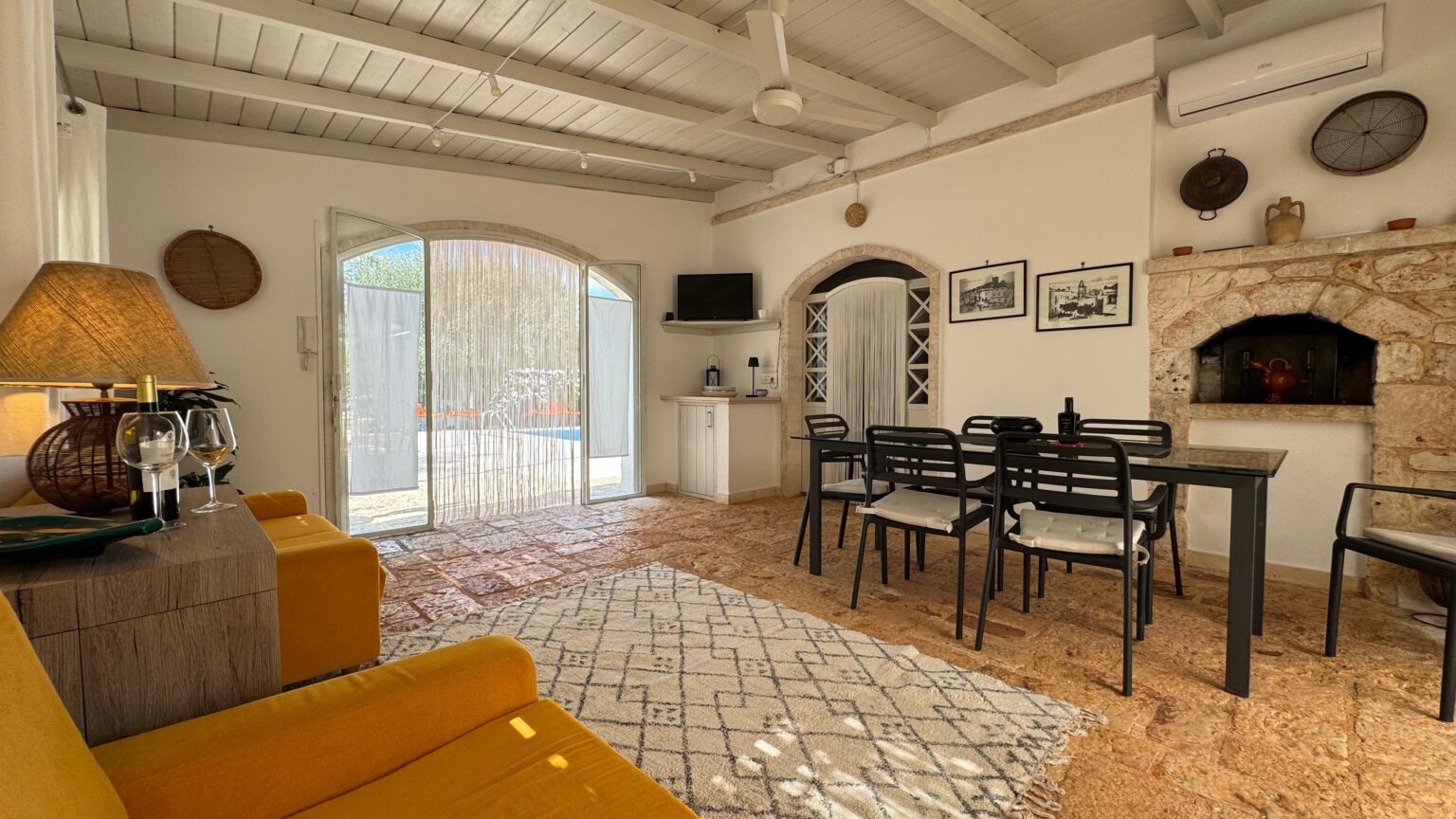 Cozy living room featuring a yellow sofa, dining table with black chairs, and a rustic fireplace, illuminated by natural light from the arched doorway. The space is decorated with framed artwork and has a warm, inviting atmosphere. | Only on SopranoVillas