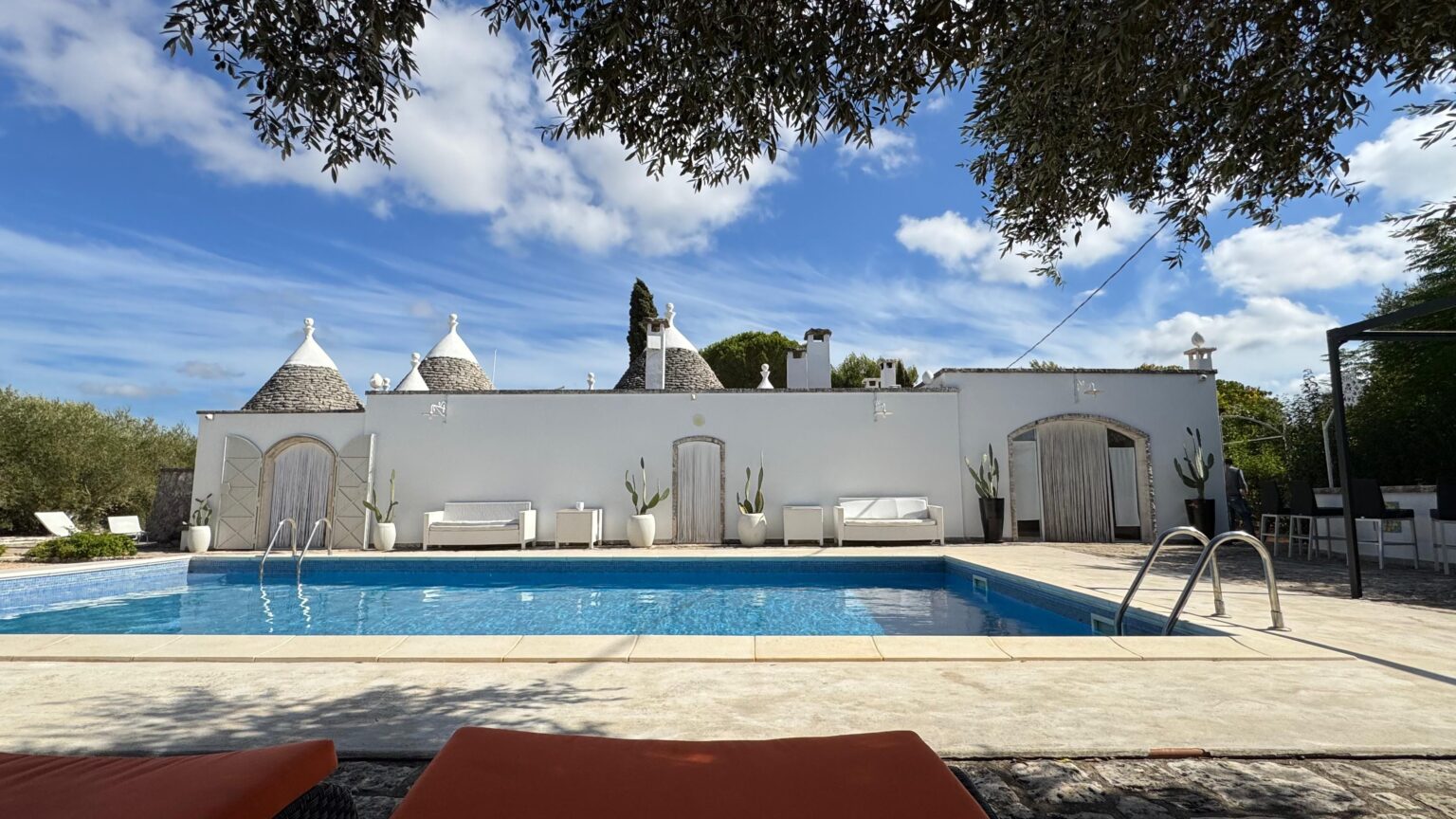Tranquil poolside view featuring a stylish outdoor space with white seating, traditional trulli architecture, and lush greenery under a clear blue sky. Ideal for relaxation and summer getaways. | Only on SopranoVillas