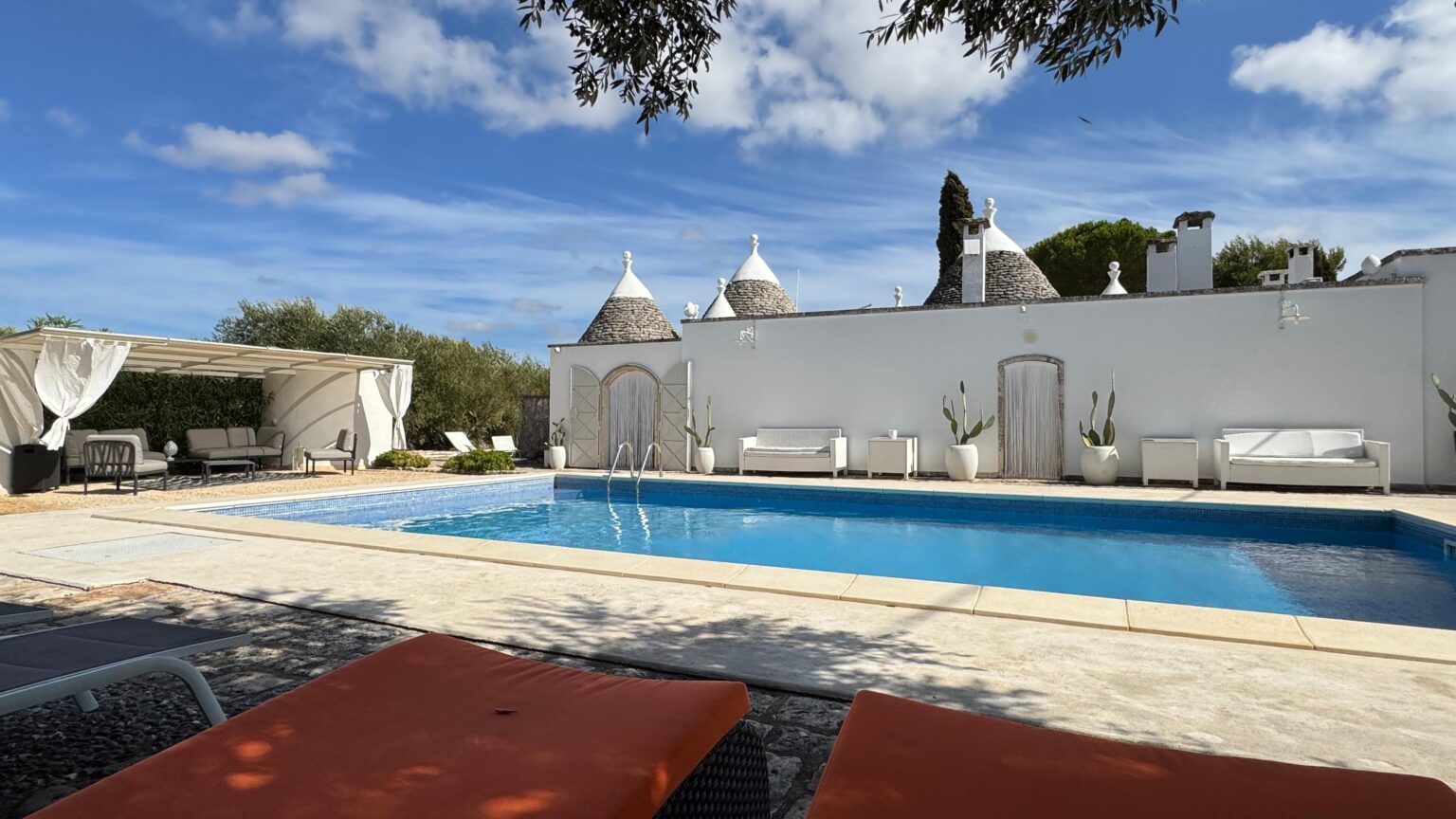 Tranquil outdoor pool area featuring modern lounge chairs, surrounded by traditional trullo architecture under a clear blue sky. Ideal for relaxation and luxury vacations. | Only on SopranoVillas